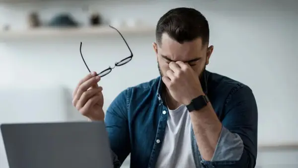 Homme fatigué, lunettes à la main, se frottant les yeux devant un ordinateur. Il est temps de penser à comment protéger ses yeux des écrans.