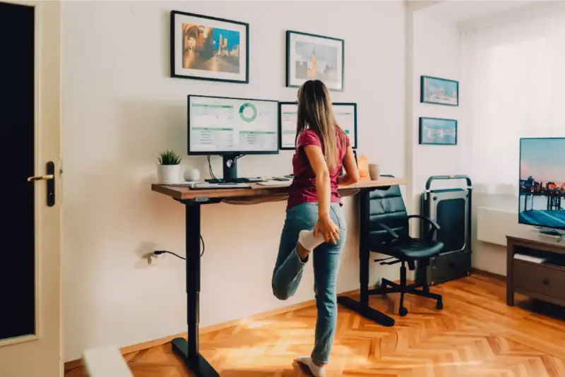 Femme s'étirant près de son bureau réglable en hauteur, pour un confort au bureau optimal. Deux écrans affichent des graphiques.
